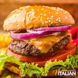 closeup of a stovetop burgers with cheese, onions, tomatoes and lettuce on a soft bun