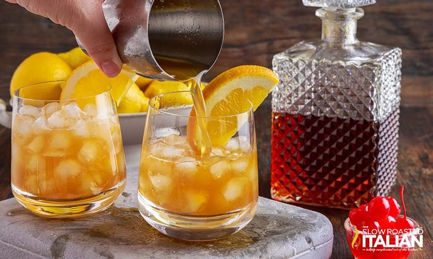 pouring amaretto sour drink from cocktail shaker into glass with ice and orange slice