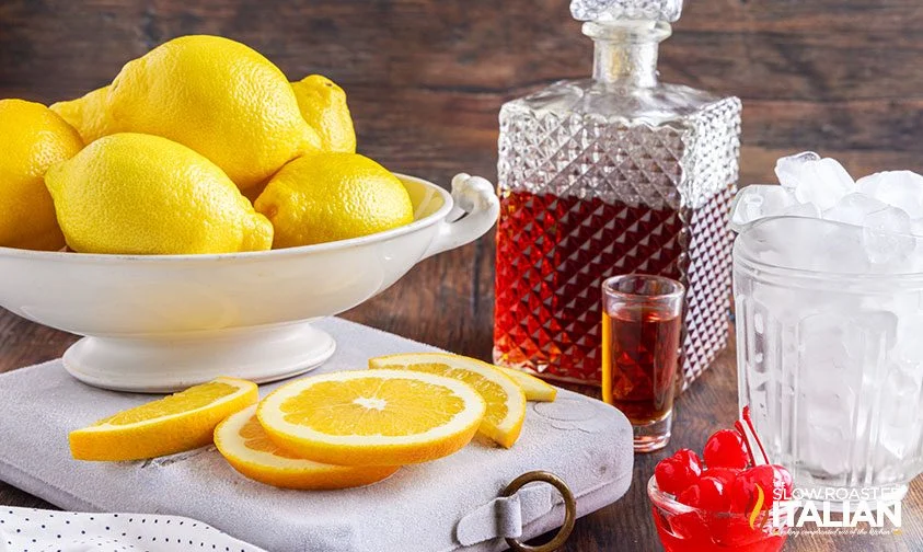 bowl of limes on cutting board with orange slices, next to decanter of amaretto and glass full of ice