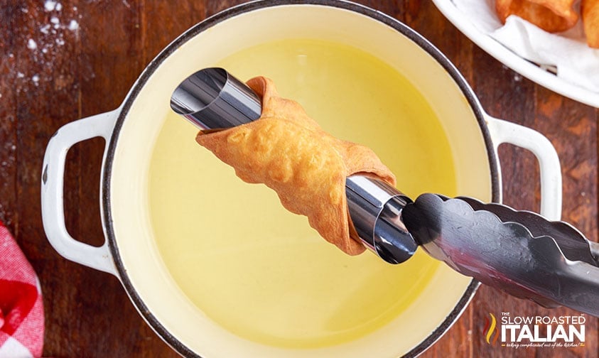lifting fried cannoli shell out of pot of oil with tongs