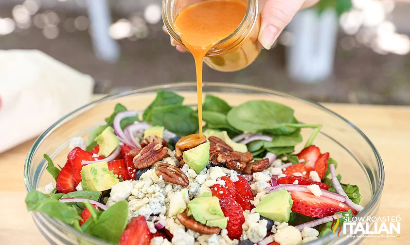 pouring homemade vinaigrette over spinach salad with berries, avocado, and candied pecans