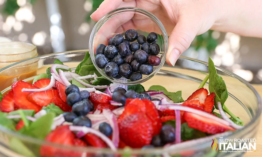 pouring blueberries into bowl with spinach, strawberries, and red onions