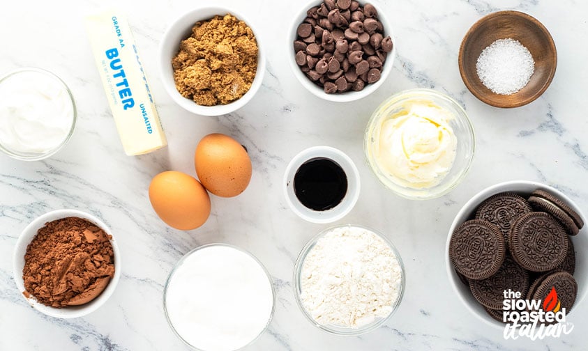 Ingredients for Oreo cheesecake brownies including butter, eggs, cocoa powder, brown sugar, chocolate chips, cream cheese, and whole Oreo cookies
