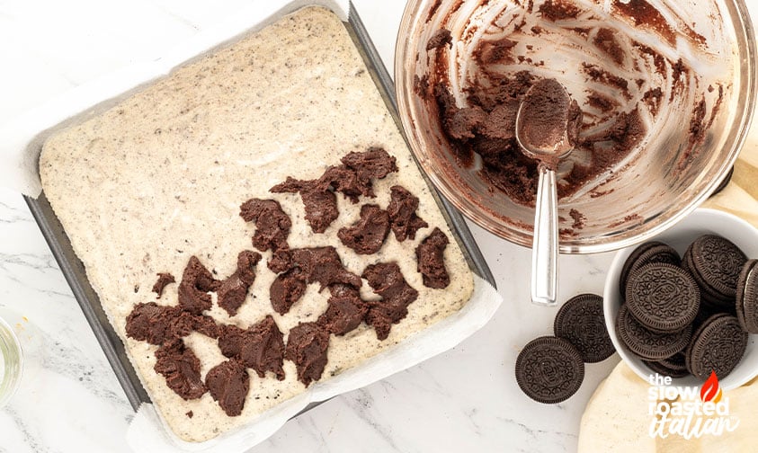 Dollops of brownie batter spooned over Oreo cheesecake filling in a lined baking pan, ready to swirl for Oreo cheesecake brownies