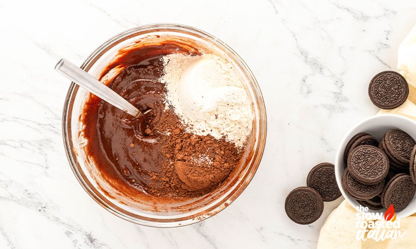 Mixing flour and cocoa powder into wet brownie batter for Oreo cheesecake brownies, with Oreo cookies on the side