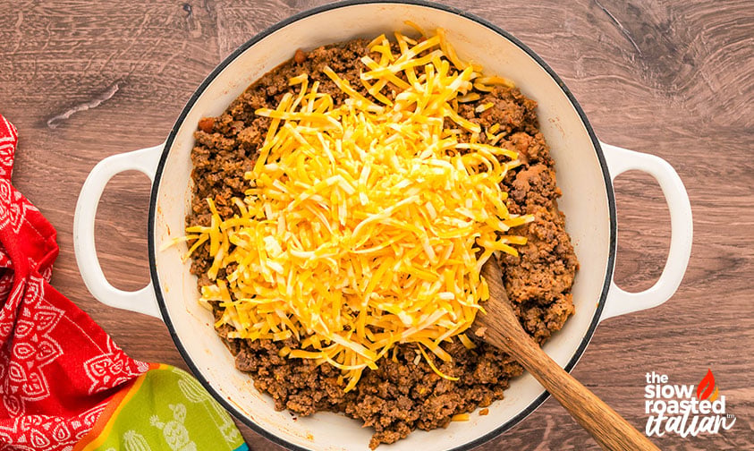 Shredded cheese added on top of cooked ground beef in a pot for refried bean casserole