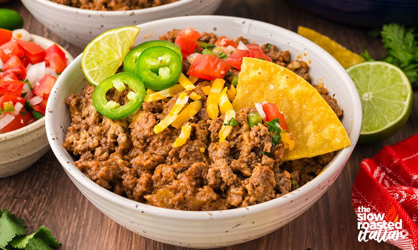 Bowl of refried bean casserole topped with jalapeños, pico de gallo, cheese, and a tortilla chip, served with lime and cilantro on the side