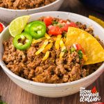 Close-up of refried bean casserole in a bowl garnished with shredded cheese, lime, jalapeño slices, pico de gallo, and a tortilla chip