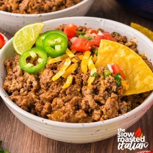Close-up of refried bean casserole in a bowl garnished with shredded cheese, lime, jalapeño slices, pico de gallo, and a tortilla chip