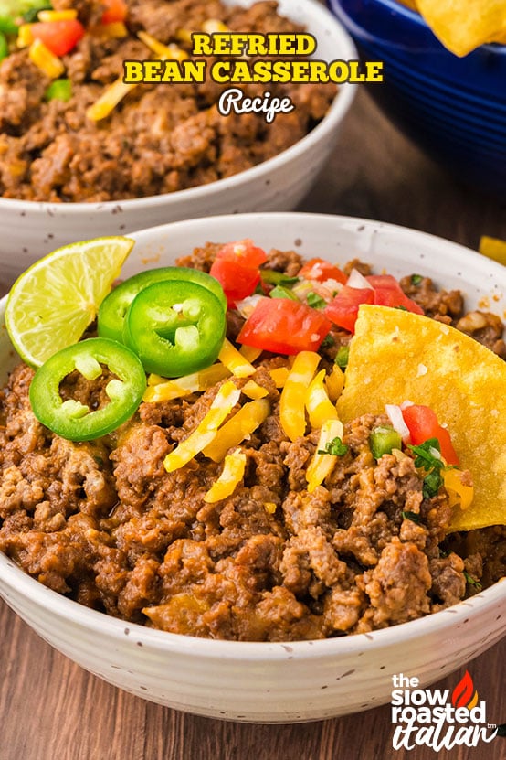 Bowl of refried bean casserole topped with shredded cheese, pico de gallo, jalapeños, lime, and a tortilla chip