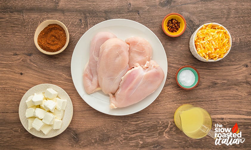 Ingredients for crockpot Mexican chicken including raw chicken breasts, cream cheese, shredded cheddar cheese, taco seasoning, red pepper flakes, salt, and chicken broth on a wooden table
