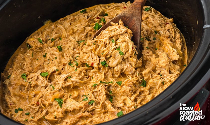 Creamy crockpot Mexican chicken shredded in the slow cooker, garnished with fresh cilantro and being scooped with a wooden spoon