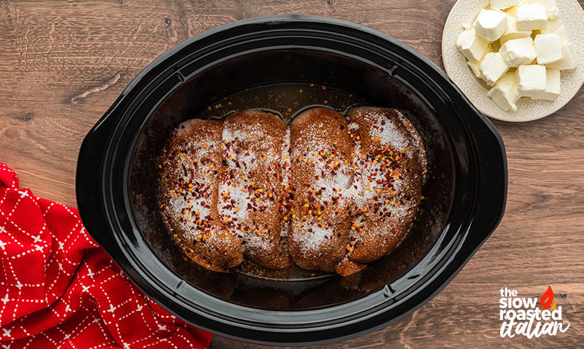 Seasoned chicken breasts in a slow cooker topped with salt and red pepper flakes, with cream cheese cubes ready to add for crockpot Mexican chicken