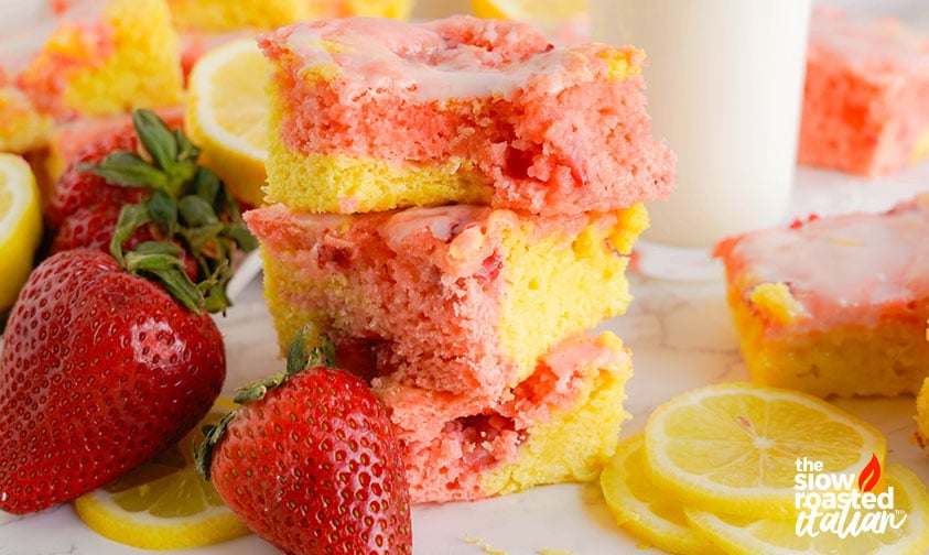 Stacked strawberry brownies with pink and yellow swirls and white glaze, surrounded by fresh strawberries and lemon slices with a glass of milk