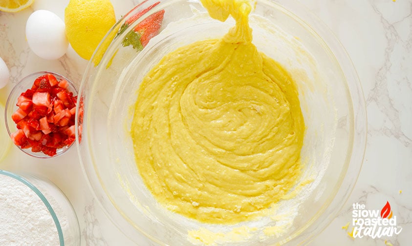 Yellow lemon batter mixed in a glass bowl for strawberry brownies, with diced strawberries and eggs on the side