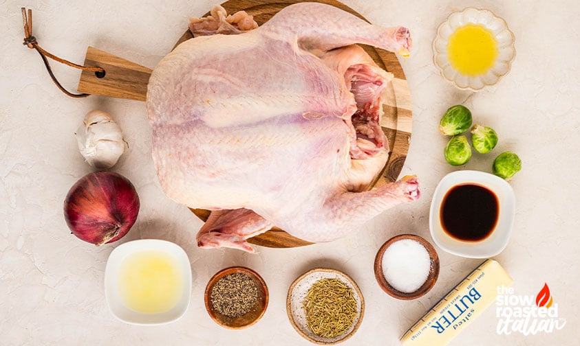 Ingredients for a roast chicken recipe including whole raw chicken, butter, garlic, red onion, Brussels sprouts, balsamic vinegar, and rosemary