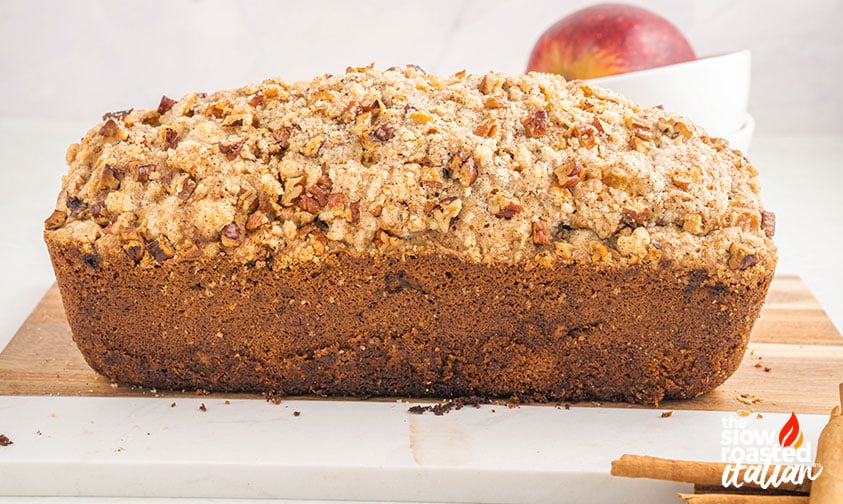 Whole baked apple bread loaf topped with pecan streusel on a wooden board, with a red apple and cinnamon sticks in the background