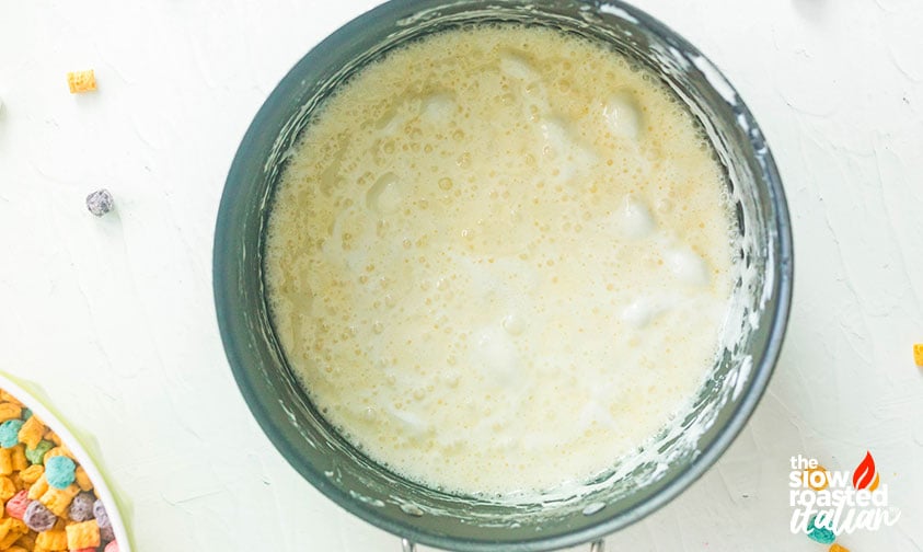 Overhead view of melted butter and marshmallow mixture bubbling in a pot, with a bowl of Cap’n Crunch cereal nearby