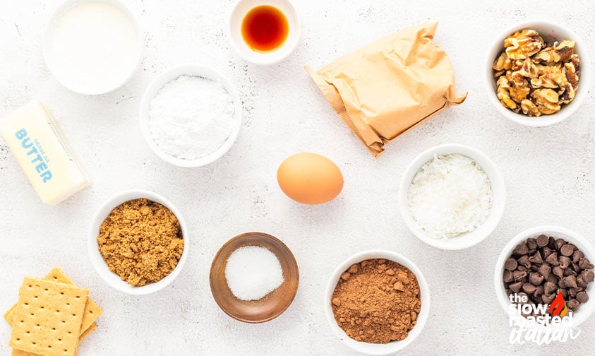 Overhead view of Nanaimo bar ingredients including butter, milk, powdered sugar, cocoa powder, coconut, walnuts, chocolate chips, graham crackers, brown sugar, egg, vanilla, and salt