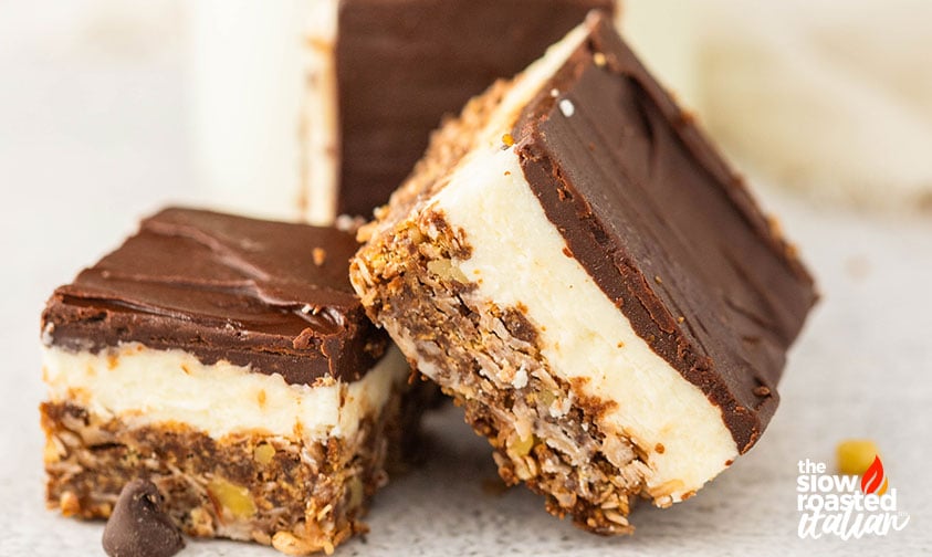 Three Nanaimo bars leaning together showing chocolate tops, creamy filling, and nutty crust layers, with chocolate chips scattered nearby