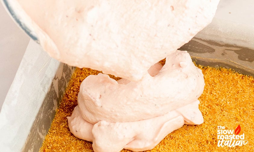 Pouring strawberry cheesecake filling over a graham cracker crust to make no bake strawberry cheesecake