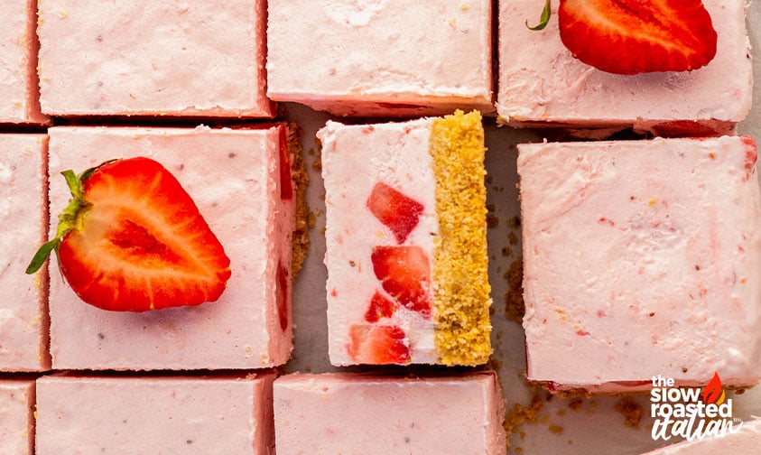 Overhead view of no bake strawberry cheesecake bars cut into squares, with one turned on its side showing the graham cracker crust and strawberry filling