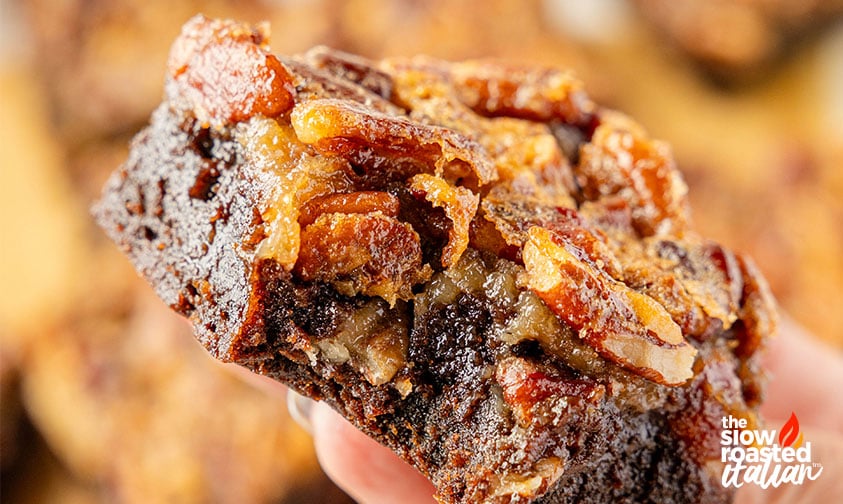Close-up of a hand holding a pecan pie brownie with a gooey pecan topping and fudgy chocolate layer