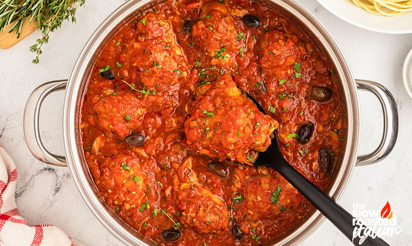 Overhead view of chicken cacciatore in a pot with a spoon lifting a piece, garnished with herbs and olives