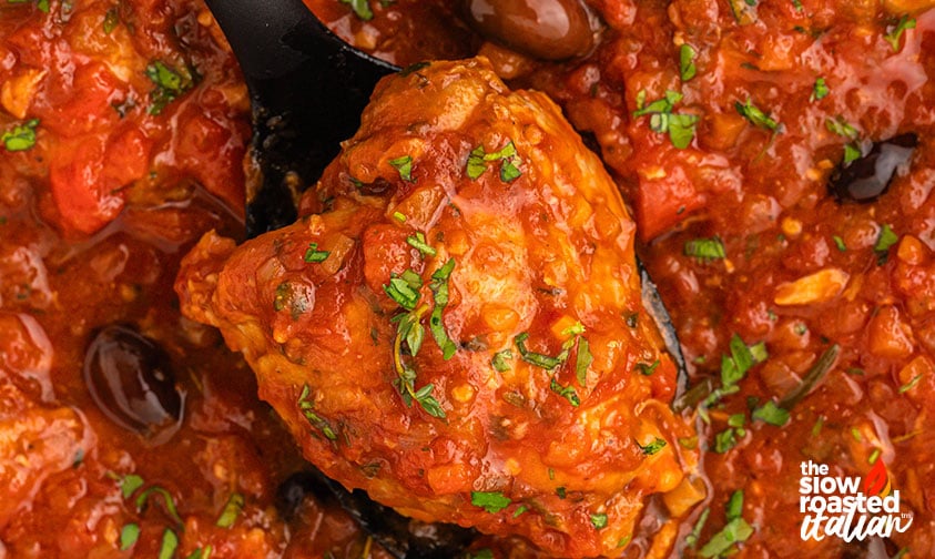 Close-up of a chicken cacciatore thigh lifted with a spoon, topped with fresh herbs and surrounded by tomato sauce and olives