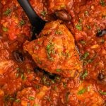Overhead close-up of chicken cacciatore being lifted with a spoon, showing tomato sauce, olives, and fresh herbs