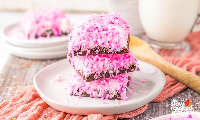 Three stacked coconut brownies with pink shredded coconut topping on a small plate, served with a glass of milk