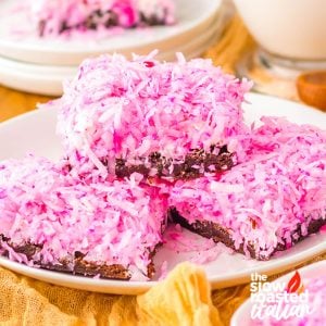 Coconut brownies topped with pink shredded coconut, stacked on a white plate