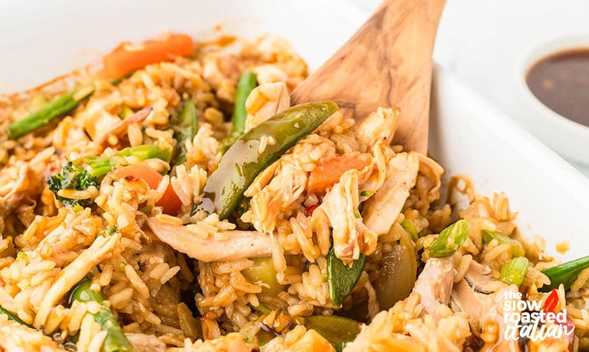 Close-up of teriyaki chicken casserole with rice, snap peas, and carrots being scooped with a wooden spoon, served with extra sauce